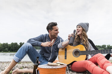 Glückliches Paar mit Bierflasche, Gitarre und Grill, das sich am Flussufer entspannt - UUF14507