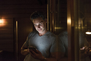 Mann lehnt am Fenster in einem dunkel beleuchteten Raum und benutzt ein Mobiltelefon - UUF14553