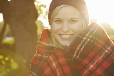 Portrait of young woman in park, tartan picnic blanket around shoulders - CUF40531