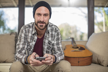 Porträt eines lächelnden jungen Mannes zu Hause auf der Couch sitzend mit Gitarre und Handy - ZEF15815