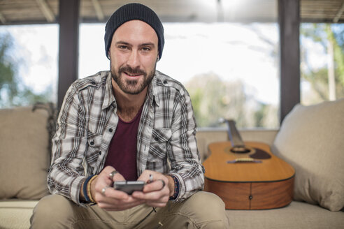 Porträt eines lächelnden jungen Mannes zu Hause auf der Couch sitzend mit Gitarre und Handy - ZEF15815