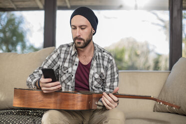 Junger Mann zu Hause auf der Couch sitzend mit Gitarre und Mobiltelefon - ZEF15816