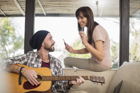 Junger Mann sitzt zu Hause auf der Couch und spielt Gitarre für eine Frau, lizenzfreies Stockfoto