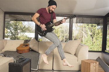 Leidenschaftlicher junger Mann spielt E-Gitarre im Wohnzimmer zu Hause und springt über einen Hügel - ZEF15832