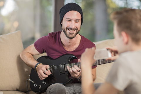 Junger Mann sitzt zu Hause auf der Couch und spielt Gitarre für einen Teenager, der ein Handyfoto macht, lizenzfreies Stockfoto