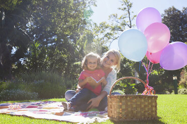 Mutter und Tochter sitzen mit Buch und Luftballons auf einer Decke im Garten - CUF41291