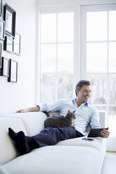 Man relaxing on sofa using digital tablet with cat - CUF41973