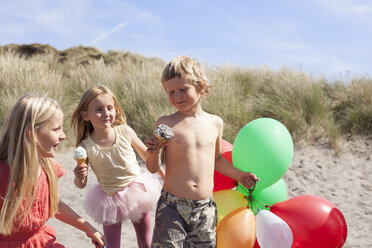 Three children eating ice creams on beach with balloons, Wales, UK - CUF42711