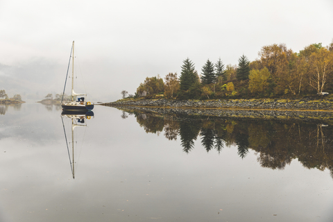 UK, Schottland, Segelboot und Baumreflexionen auf einem See im Hochland, lizenzfreies Stockfoto
