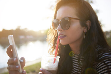 Stylish young woman with cell phone and takeaway drink outdoors at sunset - ABIF00693