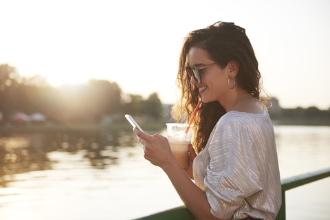 Lächelnde junge Frau mit Handy und Getränk zum Mitnehmen am Flussufer bei Sonnenuntergang, lizenzfreies Stockfoto