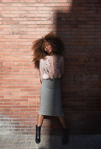 Porträt einer schönen jungen Frau mit Afrofrisur an einer Backsteinmauer im Sonnenschein, lizenzfreies Stockfoto