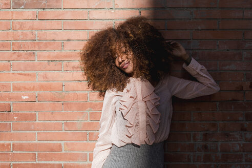 Porträt einer schönen jungen Frau mit Afrofrisur an einer Backsteinmauer im Sonnenschein - MAUF01495