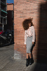 Lächelnde junge Frau mit Afrofrisur an einer Backsteinmauer in der Stadt stehend - MAUF01497