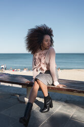 Schöne junge Frau mit Afrofrisur sitzt auf einer Bank am Strand - MAUF01502