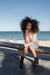 Porträt einer schönen jungen Frau mit Afrofrisur, die auf einer Bank am Strand sitzt - MAUF01503