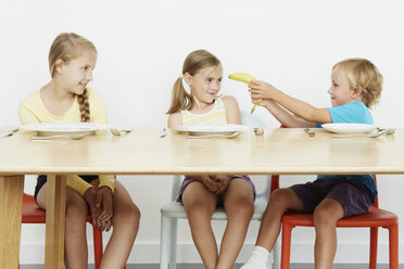 Three children at table, boy holding banana - CUF42959