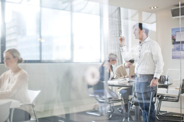 Man writing on glass wall, colleagues in background - CUF43037