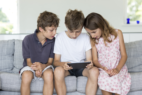 Brüder und Schwester auf dem Sofa mit Blick auf ein digitales Tablet, lizenzfreies Stockfoto