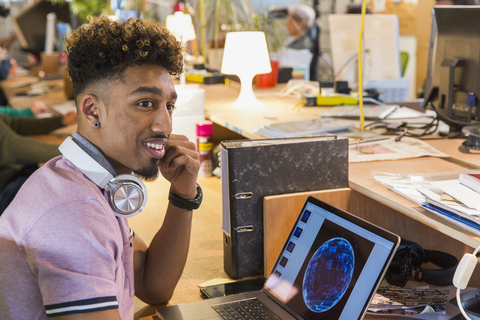 Kreativer Geschäftsmann mit Kopfhörern bei der Arbeit am Laptop im Büro, lizenzfreies Stockfoto