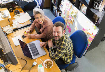Portrait lächelnd, zuversichtlich, kreative Geschäftsleute essen Müsli, arbeiten am Laptop im Büro - CAIF21067