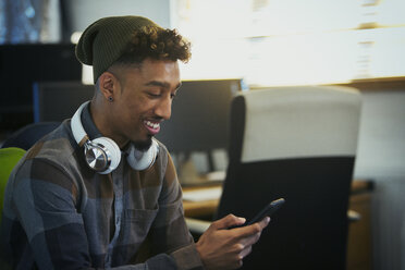 Lächelnder kreativer Geschäftsmann mit Kopfhörern und Smartphone im Büro - CAIF21076