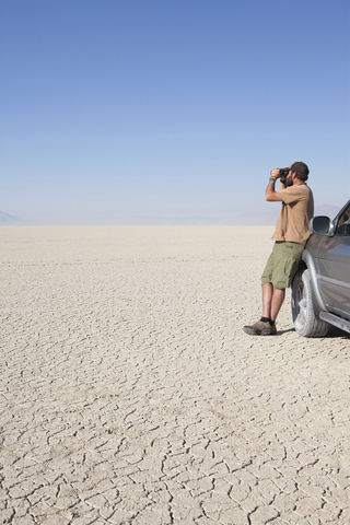 Ein Mann steht in einer trockenen Wüste, schaut durch ein Fernglas und lehnt sich an einen Lastwagen, lizenzfreies Stockfoto