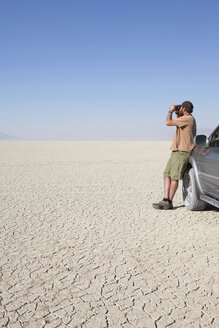 Ein Mann steht in einer trockenen Wüste, schaut durch ein Fernglas und lehnt sich an einen Lastwagen - MINF00072