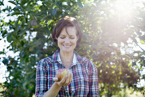 Eine Frau in einem karierten Hemd hält einen großen, frisch gepflückten Apfel im Obstgarten eines Bio-Obsthofs. - MINF00075