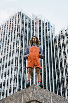 Beautiful woman wearing dungarees, standing on ledge in front of modern high-rise building - KKAF01218