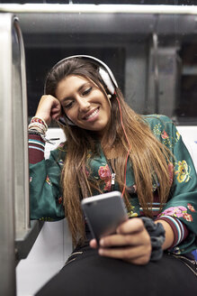 Porträt einer lächelnden Frau mit Kopfhörern, die in einem U-Bahn-Zug auf ihr Mobiltelefon schaut - JNDF00014