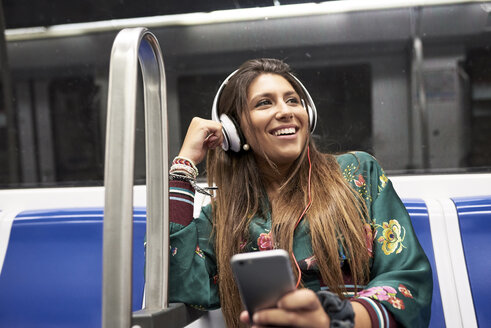 Porträt einer glücklichen Frau, die mit Kopfhörern und Smartphone in einem U-Bahn-Zug Musik hört - JNDF00015