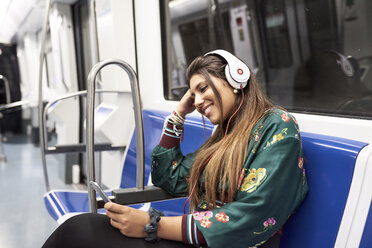 Smiling young woman listening music with headphones and smartphone in underground train - JNDF00016