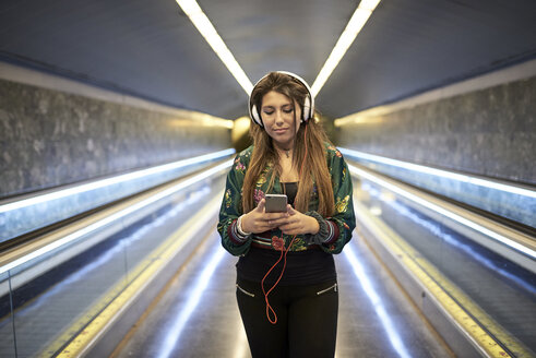Porträt einer Frau mit Kopfhörern, die in einer U-Bahn-Station auf ihr Smartphone schaut - JNDF00018