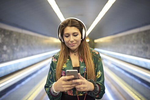 Porträt einer Frau mit Kopfhörern, die in einer U-Bahn-Station auf ihr Mobiltelefon schaut - JNDF00019