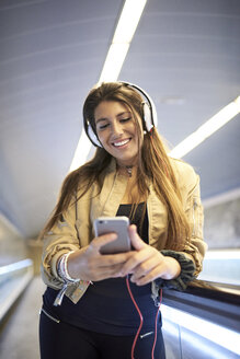 Porträt einer lächelnden jungen Frau mit Kopfhörern, die auf einer Rolltreppe steht und auf ihr Mobiltelefon schaut - JNDF00020