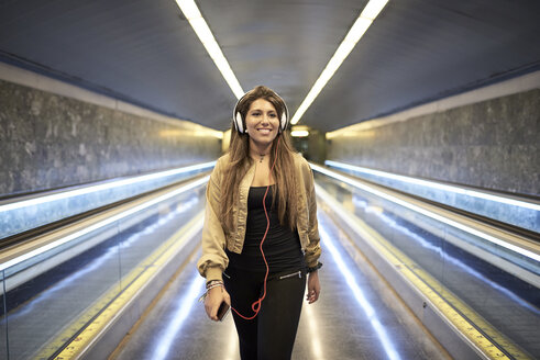 Porträt einer lächelnden Frau mit Kopfhörern und Smartphone in einer U-Bahn-Station - JNDF00021