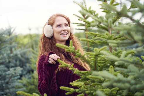 Porträt einer lächelnden rothaarigen jungen Frau mit Ohrenschützern, die einen Weihnachtsbaum in einer Baumschule aussucht - ABIF00706