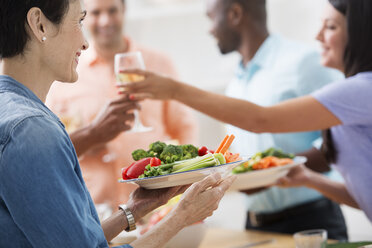 An open plan office in New York City. A working lunch, a salad buffet. A group of men and women of mixed ages and ethnicities meeting together. A woman holding out a glass of wine, and a plate of food. - MINF00098