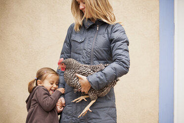 Eine Frau in einem grauen Mantel hält ein schwarz-weißes Huhn mit einem roten Hahnenkamm unter einem Arm, daneben ein junges Mädchen, das die andere Hand hält - MINF00183