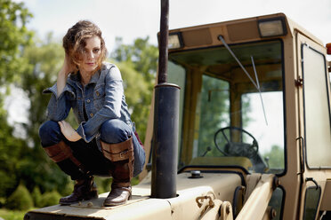 Eine junge Frau in Jeansjacke und Stiefeln auf der Motorhaube eines Traktors. - MINF00195