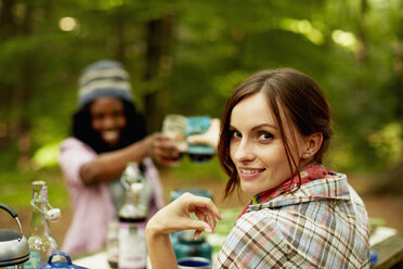 Zwei junge Frauen stoßen bei einem Picknick oder auf einem Campingplatz im Wald an. - MINF00244