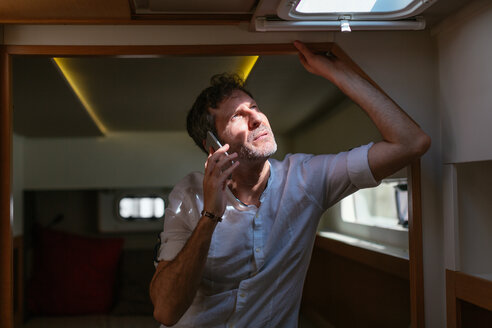 Mature man standing in catamaran cabin, looking up , using smartphone - EBSF02624