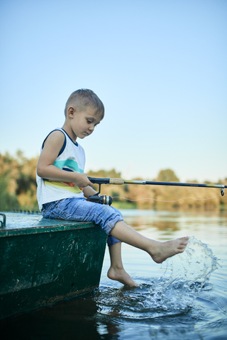 Little boy with fishing rod sitting on boat splashing with water stock photo