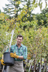 Ein Mann arbeitet in einer Bio-Gärtnerei und trägt einen jungen Baum in einem Topf. - MINF00324