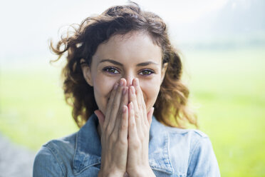 A young woman in a rural landscape, with windblown curly hair. Covering her face with her hands, and laughing. - MINF00380