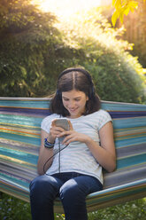 Smiling girl with headphones sitting in hammock looking at smartphone - LVF07320