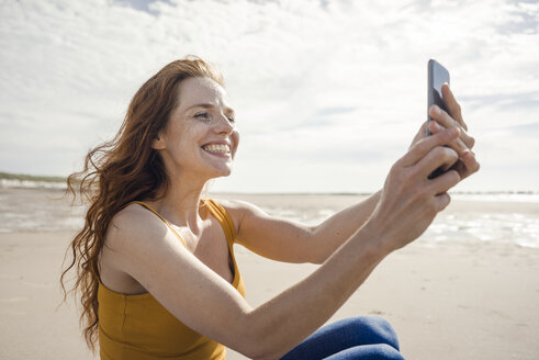 Niederlande, Zeeland, grinsende rothaarige Frau macht Selfie mit Smartphone am Strand - KNSF04194