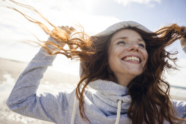Netherlands, Zeeland, portrait of happy redheaded woman wearing hooded jacket on the beach - KNSF04196