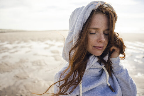 Netherlands, Zeeland, portrait of redheaded woman with freckles wearing hooded jacket on the beach - KNSF04197